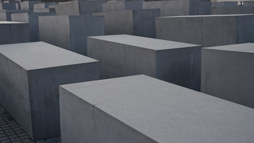 Large, rectangular concrete blocks arranged in a grid pattern, forming an abstract architectural landscape. The blocks are evenly placed on a paved ground, creating a sense of uniformity and structure.