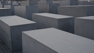 Large, rectangular concrete blocks arranged in a grid pattern, forming an abstract architectural landscape. The blocks are evenly placed on a paved ground, creating a sense of uniformity and structure.
