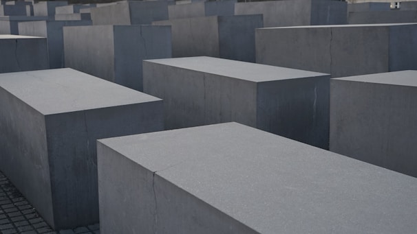 Large, rectangular concrete blocks arranged in a grid pattern, forming an abstract architectural landscape. The blocks are evenly placed on a paved ground, creating a sense of uniformity and structure.