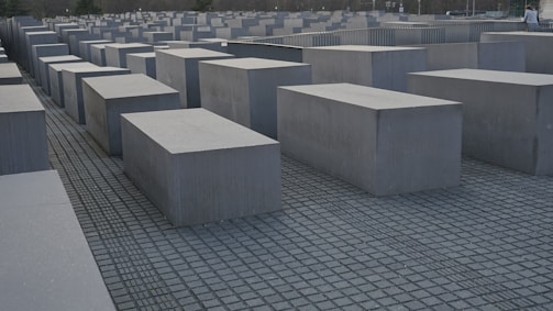 A series of large, rectangular concrete slabs are arranged in a grid pattern on a flat ground, creating a somber and structured aesthetic. The composition suggests a sense of repetition and uniformity.
