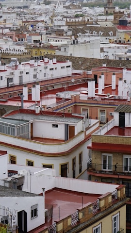 A densely packed urban neighborhood filled with various buildings of differing heights and architectural styles. Each building has a flat roof, some with antennas and satellite dishes. The structures are predominantly white with red accents, and some buildings have balconies with railings. The scene appears to be overcast, creating a somewhat muted tone for the entire area.