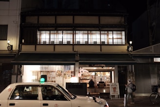A traditional Japanese building is illuminated at night. The ground floor appears to be a cozy restaurant or cafe with people visible inside. A white taxi is passing by in the foreground, while a person walks along the sidewalk. The upper floor features large windows with wooden frames.