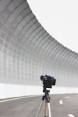 A camera is mounted on a tripod, positioned on an empty, curved road. The road is lined with a tall, curving, mesh-covered barrier, creating a tunnel-like effect. The perspective highlights the camera as the main focus against the expansive, industrial background.