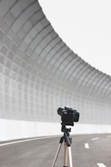 A camera is mounted on a tripod, positioned on an empty, curved road. The road is lined with a tall, curving, mesh-covered barrier, creating a tunnel-like effect. The perspective highlights the camera as the main focus against the expansive, industrial background.