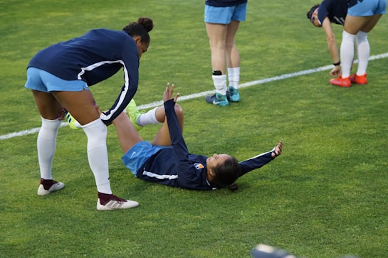A group of athletes in sportswear are on a green soccer field. Two individuals are interacting closely; one helps the other who is lying on the grass. Nearby, other athletes are stretching.