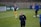 A person stands on a soccer field, wearing a navy sports jacket with a logo. In the background, another person in sports gear, including yellow shorts, is visible, holding goalkeeper gloves. A third person is near some soccer balls, facing away. The setting appears to be a practice session with advertising boards visible along the sidelines.
