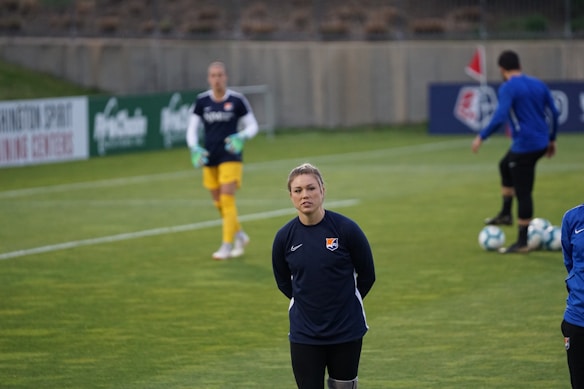 A person stands on a soccer field, wearing a navy sports jacket with a logo. In the background, another person in sports gear, including yellow shorts, is visible, holding goalkeeper gloves. A third person is near some soccer balls, facing away. The setting appears to be a practice session with advertising boards visible along the sidelines.