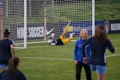 A soccer goalkeeper is making a save during a practice session, capturing the ball near the goalpost. The goalie is wearing yellow shorts and equipment, while a few other players in blue uniforms are walking on the field. Advertisements and team banners are displayed on the perimeter fencing.