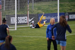 A soccer goalkeeper is making a save during a practice session, capturing the ball near the goalpost. The goalie is wearing yellow shorts and equipment, while a few other players in blue uniforms are walking on the field. Advertisements and team banners are displayed on the perimeter fencing.