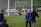 A soccer goalkeeper is making a save during a practice session, capturing the ball near the goalpost. The goalie is wearing yellow shorts and equipment, while a few other players in blue uniforms are walking on the field. Advertisements and team banners are displayed on the perimeter fencing.