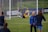 A soccer goalkeeper is making a save during a practice session, capturing the ball near the goalpost. The goalie is wearing yellow shorts and equipment, while a few other players in blue uniforms are walking on the field. Advertisements and team banners are displayed on the perimeter fencing.