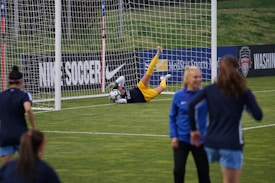 A soccer goalkeeper is making a save during a practice session, capturing the ball near the goalpost. The goalie is wearing yellow shorts and equipment, while a few other players in blue uniforms are walking on the field. Advertisements and team banners are displayed on the perimeter fencing.