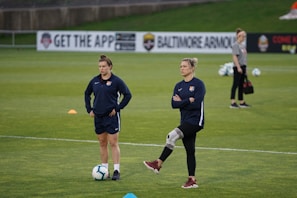 Two individuals are standing on a grassy sports field, each wearing dark athletic clothing with a logo. One person is balancing on one leg with a brace on their knee, while the other stands with hands on hips. Soccer balls and markers are visible in the background, along with a person collecting equipment.