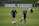 Two individuals are standing on a grassy sports field, each wearing dark athletic clothing with a logo. One person is balancing on one leg with a brace on their knee, while the other stands with hands on hips. Soccer balls and markers are visible in the background, along with a person collecting equipment.