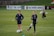Two individuals are standing on a grassy sports field, each wearing dark athletic clothing with a logo. One person is balancing on one leg with a brace on their knee, while the other stands with hands on hips. Soccer balls and markers are visible in the background, along with a person collecting equipment.