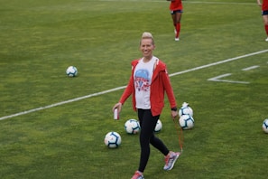 A person in athletic clothing walks across a grass field, holding a drink can and a skipping rope. Several soccer balls are scattered on the field. Other individuals in red athletic attire are in the background, suggesting a sports practice or event.