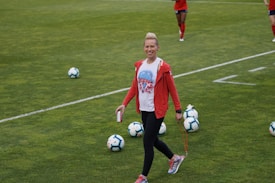 A person in athletic clothing walks across a grass field, holding a drink can and a skipping rope. Several soccer balls are scattered on the field. Other individuals in red athletic attire are in the background, suggesting a sports practice or event.