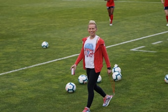 A person in athletic clothing walks across a grass field, holding a drink can and a skipping rope. Several soccer balls are scattered on the field. Other individuals in red athletic attire are in the background, suggesting a sports practice or event.