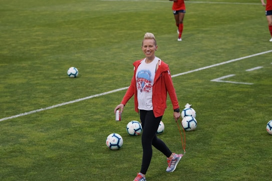 A person in athletic clothing walks across a grass field, holding a drink can and a skipping rope. Several soccer balls are scattered on the field. Other individuals in red athletic attire are in the background, suggesting a sports practice or event.