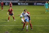 Two soccer players are in the middle of a play. One player in a white and blue uniform appears to be controlling the ball while the other player in a red and black uniform is closely defending. There are other players on the field, with a goalkeeper in a turquoise uniform in the background.