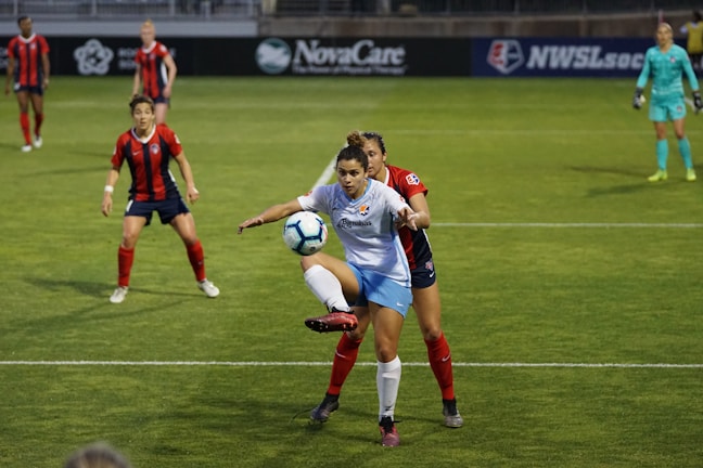 Two soccer players are in the middle of a play. One player in a white and blue uniform appears to be controlling the ball while the other player in a red and black uniform is closely defending. There are other players on the field, with a goalkeeper in a turquoise uniform in the background.