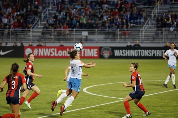 A women's soccer match is taking place on a well-maintained grass field. Players from two teams, one in red and black and the other in blue and white, are actively engaged in the game. One player is jumping to head the ball, while others are positioned around, focused on the play. The stadium stands in the background are filled with spectators, and advertising banners are visible on the sideline.