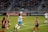 A women's soccer match is taking place on a well-maintained grass field. Players from two teams, one in red and black and the other in blue and white, are actively engaged in the game. One player is jumping to head the ball, while others are positioned around, focused on the play. The stadium stands in the background are filled with spectators, and advertising banners are visible on the sideline.