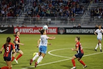 A women's soccer match is taking place on a well-maintained grass field. Players from two teams, one in red and black and the other in blue and white, are actively engaged in the game. One player is jumping to head the ball, while others are positioned around, focused on the play. The stadium stands in the background are filled with spectators, and advertising banners are visible on the sideline.