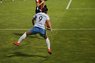A soccer player is captured in mid-air, seemingly in the midst of a dynamic move or leap on the field. The player wears a jersey with the number 9 and the name Kawasumi on the back, along with white shorts, long white socks, and bright orange cleats. The green grass of the soccer field with white boundary lines can be seen, suggesting an active sports setting. Another player in the background is partially visible, wearing a different team’s uniform.