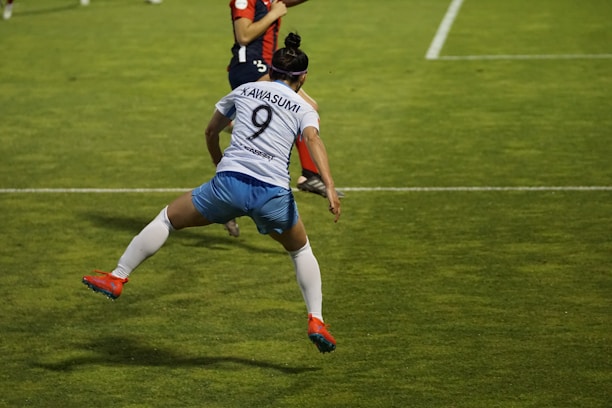 A soccer player is captured in mid-air, seemingly in the midst of a dynamic move or leap on the field. The player wears a jersey with the number 9 and the name Kawasumi on the back, along with white shorts, long white socks, and bright orange cleats. The green grass of the soccer field with white boundary lines can be seen, suggesting an active sports setting. Another player in the background is partially visible, wearing a different team’s uniform.