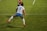 A soccer player is captured in mid-air, seemingly in the midst of a dynamic move or leap on the field. The player wears a jersey with the number 9 and the name Kawasumi on the back, along with white shorts, long white socks, and bright orange cleats. The green grass of the soccer field with white boundary lines can be seen, suggesting an active sports setting. Another player in the background is partially visible, wearing a different team’s uniform.
