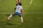 A soccer player is captured in mid-air, seemingly in the midst of a dynamic move or leap on the field. The player wears a jersey with the number 9 and the name Kawasumi on the back, along with white shorts, long white socks, and bright orange cleats. The green grass of the soccer field with white boundary lines can be seen, suggesting an active sports setting. Another player in the background is partially visible, wearing a different team’s uniform.