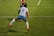 A soccer player is captured in mid-air, seemingly in the midst of a dynamic move or leap on the field. The player wears a jersey with the number 9 and the name Kawasumi on the back, along with white shorts, long white socks, and bright orange cleats. The green grass of the soccer field with white boundary lines can be seen, suggesting an active sports setting. Another player in the background is partially visible, wearing a different team’s uniform.