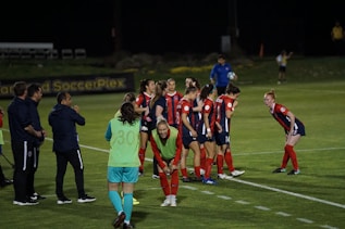 A group of female soccer players, dressed in red and navy uniforms, are gathered on the sidelines with some bending forward. A coach and staff members stand nearby, some clapping or giving instructions. The scene takes place on a well-lit soccer field at night with a banner in the background.