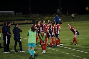 A group of female soccer players, dressed in red and navy uniforms, are gathered on the sidelines with some bending forward. A coach and staff members stand nearby, some clapping or giving instructions. The scene takes place on a well-lit soccer field at night with a banner in the background.