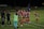 A group of female soccer players, dressed in red and navy uniforms, are gathered on the sidelines with some bending forward. A coach and staff members stand nearby, some clapping or giving instructions. The scene takes place on a well-lit soccer field at night with a banner in the background.