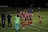 A group of female soccer players, dressed in red and navy uniforms, are gathered on the sidelines with some bending forward. A coach and staff members stand nearby, some clapping or giving instructions. The scene takes place on a well-lit soccer field at night with a banner in the background.