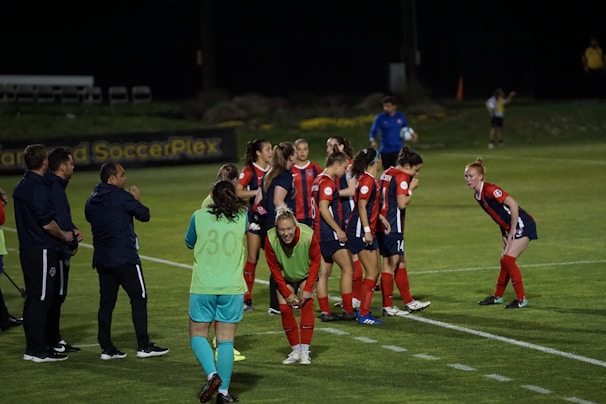 A group of female soccer players, dressed in red and navy uniforms, are gathered on the sidelines with some bending forward. A coach and staff members stand nearby, some clapping or giving instructions. The scene takes place on a well-lit soccer field at night with a banner in the background.