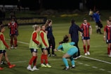 A group of female soccer players on a field, some wearing green practice vests over their red and blue uniforms. They appear to be preparing for a match or warming up. There is a mix of activity and casual interaction, with some players standing in conversation and others jogging or stretching. The soccer field has a clearly marked sideline and goal area, while the background shows a sign for a sports complex.