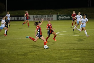 A group of female soccer players in action during a game on a grass field. One player wearing a red and navy uniform is about to kick the ball, while other players in similar uniforms and white uniforms are either running towards her or observing. The scene is set in an outdoor stadium with a sign indicating Washington Spirit Training Centers in the background.