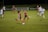 A group of female soccer players in action during a game on a grass field. One player wearing a red and navy uniform is about to kick the ball, while other players in similar uniforms and white uniforms are either running towards her or observing. The scene is set in an outdoor stadium with a sign indicating Washington Spirit Training Centers in the background.