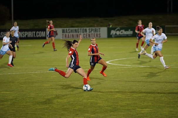 A group of female soccer players in action during a game on a grass field. One player wearing a red and navy uniform is about to kick the ball, while other players in similar uniforms and white uniforms are either running towards her or observing. The scene is set in an outdoor stadium with a sign indicating Washington Spirit Training Centers in the background.
