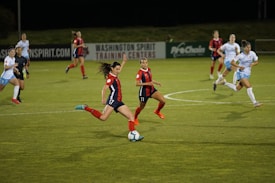 A group of female soccer players in action during a game on a grass field. One player wearing a red and navy uniform is about to kick the ball, while other players in similar uniforms and white uniforms are either running towards her or observing. The scene is set in an outdoor stadium with a sign indicating Washington Spirit Training Centers in the background.
