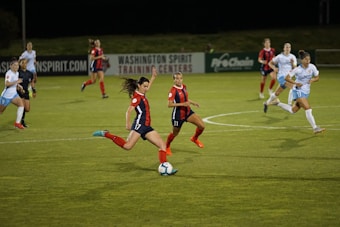 A group of female soccer players in action during a game on a grass field. One player wearing a red and navy uniform is about to kick the ball, while other players in similar uniforms and white uniforms are either running towards her or observing. The scene is set in an outdoor stadium with a sign indicating Washington Spirit Training Centers in the background.