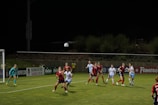 A women's soccer match is in progress. Several players wearing red and black striped jerseys and white jerseys are intensely focused on a soccer ball in mid-air. The soccer field is under bright lights against a dark night sky. One player is preparing to head the ball, while a goalkeeper in turquoise gear is positioned near the goal post.