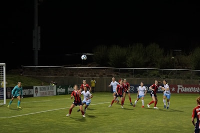 A women's soccer match is in progress. Several players wearing red and black striped jerseys and white jerseys are intensely focused on a soccer ball in mid-air. The soccer field is under bright lights against a dark night sky. One player is preparing to head the ball, while a goalkeeper in turquoise gear is positioned near the goal post.