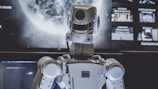 A humanoid robot is positioned in front of a large screen with a planetary image, possibly of the moon. The robot appears to be facing the screen, lit by both the screen and ambient lighting.