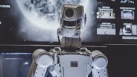 A humanoid robot is positioned in front of a large screen with a planetary image, possibly of the moon. The robot appears to be facing the screen, lit by both the screen and ambient lighting.