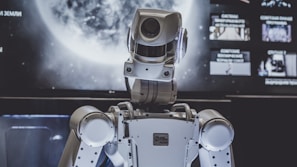 A humanoid robot is positioned in front of a large screen with a planetary image, possibly of the moon. The robot appears to be facing the screen, lit by both the screen and ambient lighting.