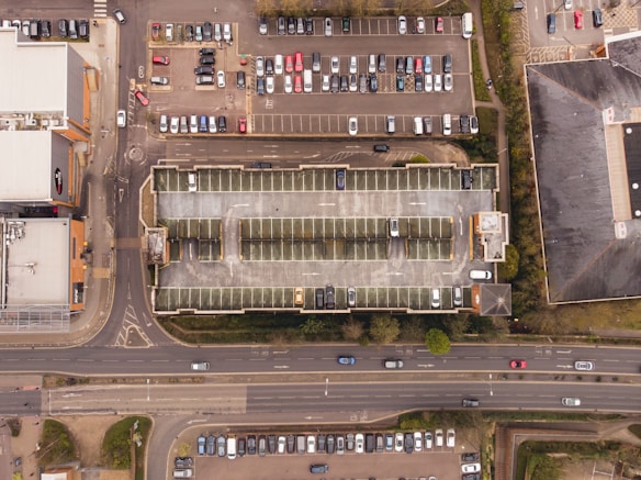 Aerial view of a multi-story parking garage surrounded by parking lots with numerous parked cars. Adjacent to the garage, a busy road runs horizontally across the bottom of the image with several moving vehicles. Surrounding the parking infrastructure are various buildings and green spaces.